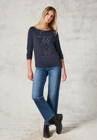 Marineblaues Langarmshirt mit einem geometrischen silbernen Grafik, kombiniert mit blauen weitem Jeans und schwarzen Schnürstiefeln auf einem strukturierten Hintergrund.