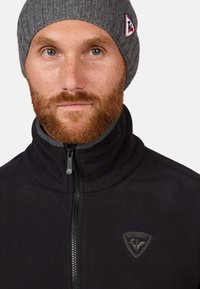 Schwarze Fleecejacke mit hohem Kragen, Frontreißverschluss und grauem Innenfutter. Ausgestattet mit einer grauen Strickmütze mit einem Logo-Patch.