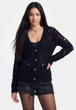 Ung kvinde med langt mørkt hår, iført en sort revet cardigan, sort tanktop, sorte revnede shorts og øreringe og halskæde i stjerneform.