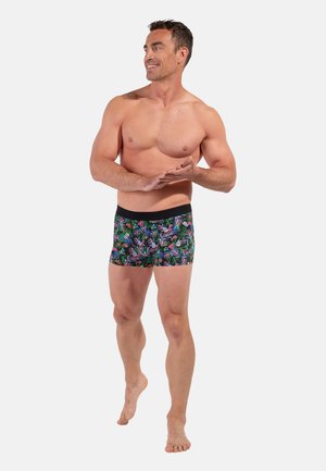 Muskularny mężczyzna boso, uśmiechający się i idący, ubrany w czarne bokserki z kolorowym tropikalnym wzorem liści i kwiatów.