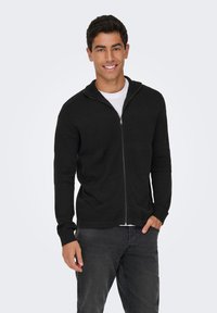 Crna zip-up cardigan s teksturiranim pletivom, visokim ovratnikom i rebrastim manžetama, uparen s bijelom majicom i tamnim trapericama.