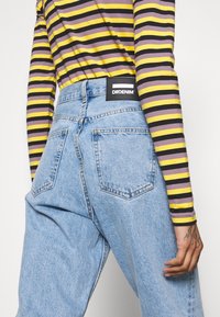 Ljusblå denimjeans med hög midja, med två bakfickor och en svart "DRDENIM"-etikett på midjebandet. Randig långärmad skjorta synlig.