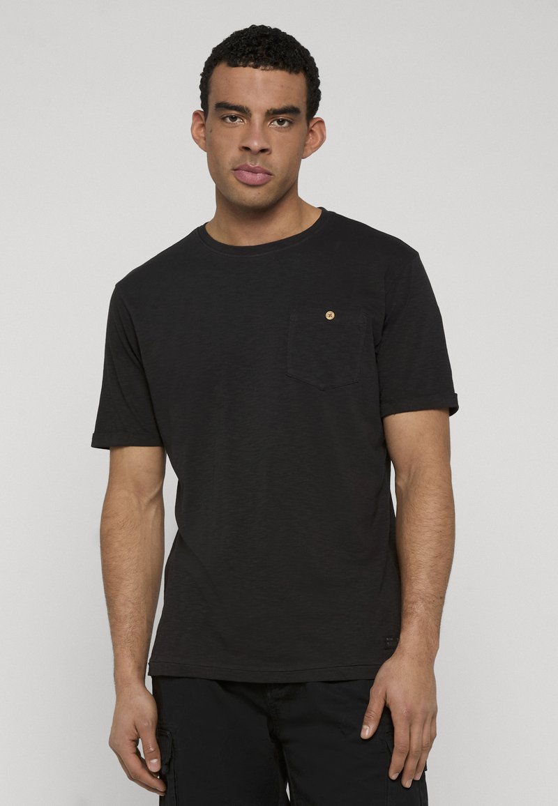 Homme aux cheveux courts et bouclés portant un t-shirt noir uni avec une petite poche poitrine, debout devant un fond gris.