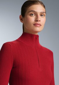 Ung kvinna med blå ögon som bär en röd ribbad polotröja med dragkedja fram, mot en enfärgad grå bakgrund.