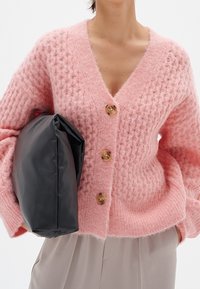 Pink knitted cardigan with a V-neck, featuring three wooden buttons. Holding a black leather bag with a soft, smooth texture.