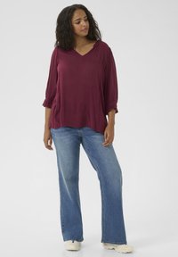Top en V de couleur bordeaux avec des manches 3/4, texture douce et poignets froncés, associé à un jean ample bleu clair et des baskets beiges.