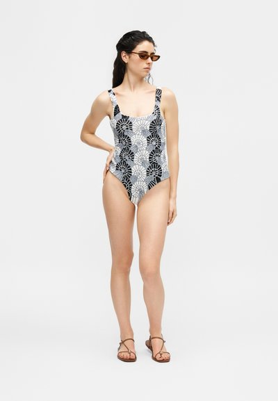 Γυναίκα που στέκεται, φοράει ολόσωμο μαγιό με μοτίβο από μαύρα και λευκά κοχύλια, καφέ δερμάτινα σανδάλια με λουράκια και καφέ γυαλιά ηλίου cat-eye.