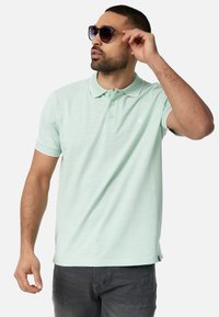 Lyst grøn polo shirt med en struktureret overflade, der har krave, to knapper og hvide accenter. Båret med mørkegrå jeans og solbriller.