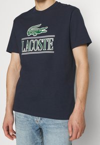 Man wearing a navy blue short-sleeve t-shirt with a green crocodile logo and "LACOSTE" text, paired with light blue jeans.