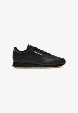 Zapatilla deportiva negra Reebok de corte bajo con suela de goma, cierre de cordones en la parte frontal y parche con el logo que muestra la bandera británica en el lateral y el talón.