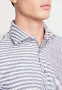 Randig skjorta med krage, med smala blå och vita ränder, grå knapp och textil med struktur. Nära vy av halsringningen.