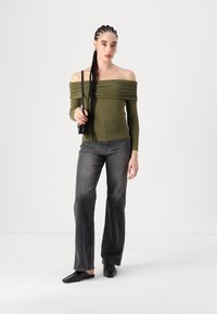Olivengrønn avskjærmet topp med samlet detaljering, kombinert med mørkegrå vide jeans. Modellen holder en liten svart veske og har på seg svarte leiligheter.