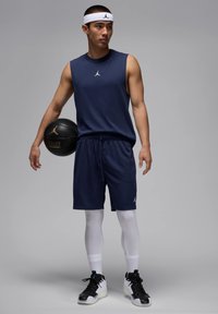 Top sin mangas de color marino, pantalones cortos a juego con detalles perforados, mallas de compresión blancas, zapatillas negras y blancas, sosteniendo un balón de baloncesto.