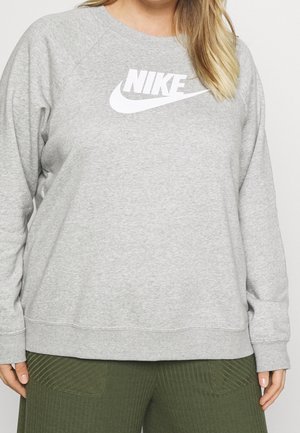 Kvinde iført en lysegrå Nike sweatshirt med hvidt logo og grønne ribstrikkede bukser, stående mod en ensfarvet baggrund.