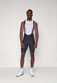 Μαύρα ποδηλατικά σορτς με τιράντες ρυθμιζόμενες, λεία υφή και εφαρμοστή σχεδίαση. Φορεμένα πάνω από λευκό κοντομάνικο τοπ.