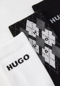 Drei Socken: eine weiße mit schwarzem "HUGO"-Text, eine schwarze mit geometrischem Muster und weißem "HUGO"-Text sowie eine strukturierte schwarze Rippsocke.