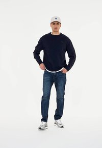 Námornícky sveter s okrúhlym výstrihom, kombinovaný s modrými džínami a striebornými teniskami. Modelka má na hlave svetlošedú čiapku s logom.