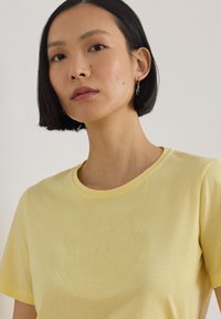 Γυναίκα με κοντά μαύρα μαλλιά φοράει χρυσά σκουλαρίκια και μια παστέλ κίτρινη μπλούζα με στρογγυλή λαιμόκοψη και διακριτικό ανάγλυφο σχέδιο στο στήθος.