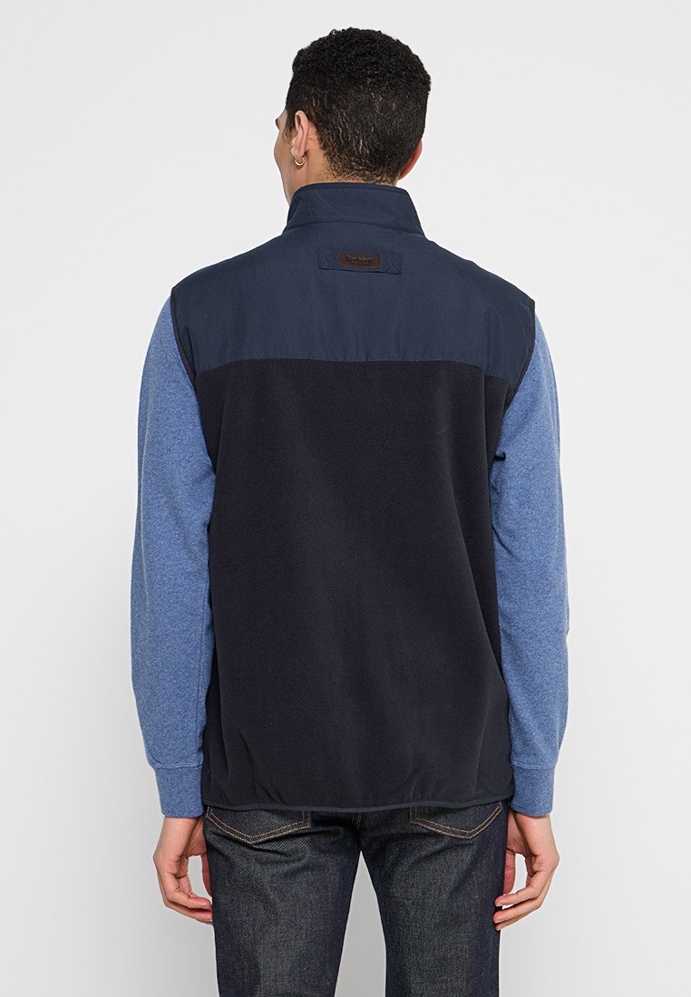 Homme portant un gilet sans manches noir et bleu marine sur une chemise à manches longues bleue, se tenant de dos avec les mains détendues le long du corps.