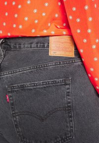 Jean noir Levi's 501 avec poche arrière, patch en cuir de la marque et étiquette rouge, associé à une chemise orange à petits motifs floraux blancs et jaunes.