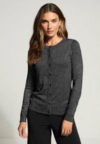 Svart, långärmad kofta med en texturerad stickad design, rund halsringning och knappstängning framtill. Bärs över svarta byxor.