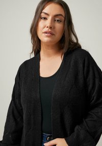 Schwarze offene Strickjacke aus texturiertem Stoff, mit lockerer Passform, Diamantmuster-Details und weitem Ärmel.