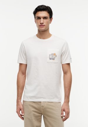 Άνδρας που φορεί λευκό κοντομάνικο μπλουζάκι με λουλουδένιο και λογότυπο σχέδιο στην τσέπη στο στήθος και μπεζ παντελόνι.