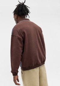 Καφέ πουλόβερ φούτερ με γιακά, που διαθέτει μπλε πλευρικές πάνελ και λευκές ραφές. Μαλακή υφή με χαλαρή εφαρμογή, κατάλληλο για στρώσεις.