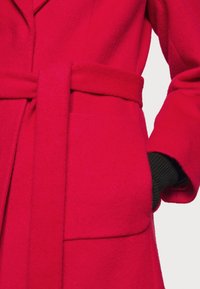 Manteau en laine rouge avec ceinture nouée à la taille et une main gantée de noir dans la poche droite.