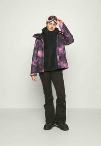 Giacca da sci con un motivo floreale viola e nero, strato di pile nero sotto, abbinata a pantaloni da sci neri e stivali da neve neri.