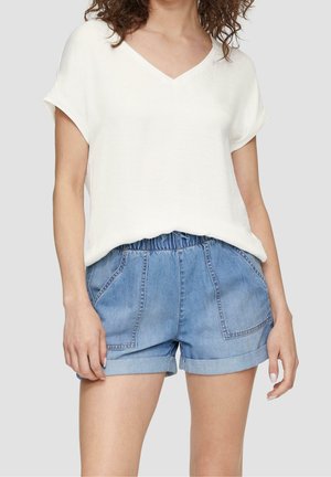 Frau trägt ein weißes V-Ausschnitt-Oberteil mit kurzen Ärmeln, das in blaue Jeans-Shorts mit elastischem Bund und umgeschlagenem Saum gesteckt ist, die vorne Taschen haben.