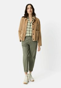 Cordjacke in Hellbraun mit schwarzen Knöpfen, grünes kariertes Hemd, olivgrüne Hose und weiße Sneaker mit Gummisohle. Lässiges Outfit mit Taschen.