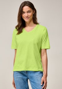 T-shirt à col V et manches courtes en vert citron brillant, fabriqué à partir d'un tissu léger. Associe à un jean à rayures bleu clair.