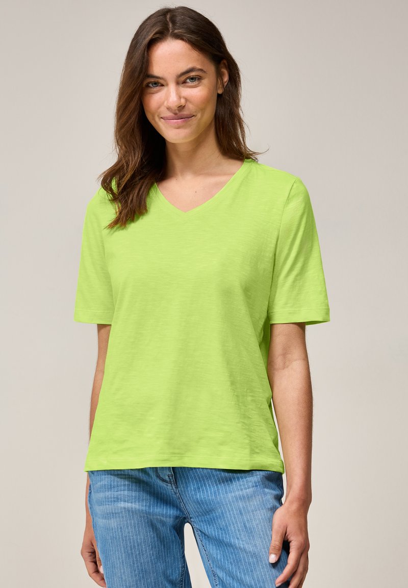 T-shirt à col V et manches courtes en vert citron brillant, fabriqué à partir d'un tissu léger. Associe à un jean à rayures bleu clair.
