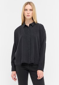 Camisa negra abotonada hecha de una tela suave, con un corte relajado, cuello puntiagudo y detalles de costura a lo largo de los hombros.