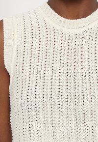 Gros plan d'un haut sans manches blanc en maille avec un col rond côtelé et un petit motif de points ouverts.