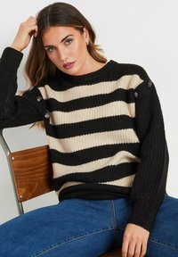 Maglione a righe nero e crema con texture a coste, caratterizzato da bottoni decorativi sulle spalle, abbinato a jeans blu.