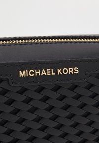 Černá tkaná peněženka s hladkým koženým obložením, zlatým kováním a viditelným zipem. Logo "MICHAEL KORS" je vyraženo na přední straně.