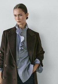 Žena s vlasy staženými dozadu má na sobě hnědý dvouřadý kabát přes modrou košili s volánkovým límcem a stojí s jednou rukou v kapse před šedou zdí.