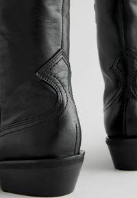 Bottes de cowboy en cuir noir avec un bout pointu, une surface texturée, des coutures décoratives et un talon épais. Design accentué sur la tige.