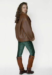 Brun læderjakke i oversized stil med syninger over grønne velourleggings, suppleret med knæhøje brune støvler med spænde-detaljer.