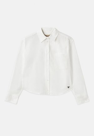 Chemise blanche à manches longues raccourcie en coton. Elle présente un devant à boutons, une poche sur la poitrine et un petit logo noir en bas à gauche.