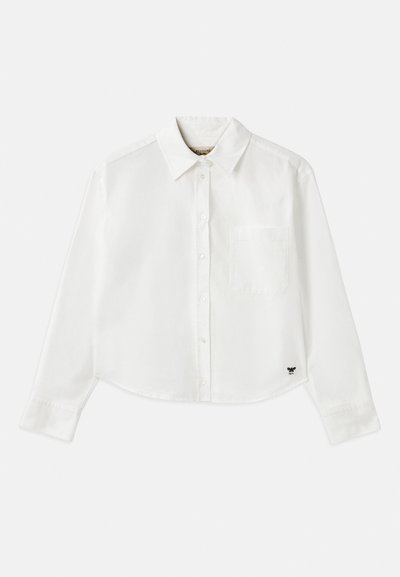 Chemise blanche à manches longues raccourcie en coton. Elle présente un devant à boutons, une poche sur la poitrine et un petit logo noir en bas à gauche.