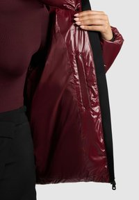 Burgunderfarbene Pufferjacke mit glänzender Innenseite, schwarzem Besatz und einer Reißverschlusstasche. Glatte Textur und mittellanges Design.