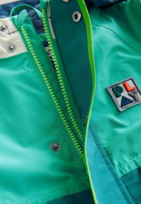 Grüne Jacke aus leichtem Material, mit einem kontrastierenden hellgrünen Reißverschluss, Druckknöpfen und einem Markenpatch auf der Brust.