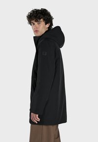 Parka negra con capucha, diseño elegante y marca visible. Presenta una textura suave, mangas largas y un corte relajado.