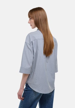 Femme aux cheveux longs et lisses portant une chemise ample rayée avec des manches aux coudes et un jean bleu, tournée vers la gauche sur un fond uni.