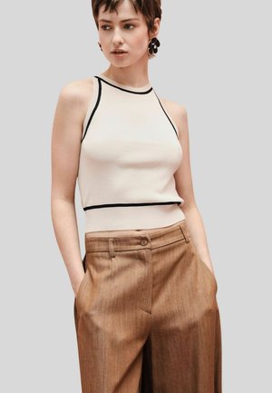 Giovane donna con capelli corti che indossa un top crema senza maniche con rifiniture nere e pantaloni marroni a vita alta, mani nelle tasche.