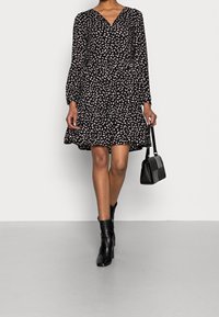 Schwarzes Kleid mit weißem Blumenmuster, V-Ausschnitt, langen Ärmeln und gestuftem Rock. Getragen mit schwarzen Knöchelboots und einer kleinen schwarzen Handtasche.