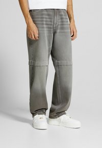 Man som bär lösa grå byxor med sömdetalj ovanför knäna och vita sneakers, stående på en jämn ljusgrå yta.